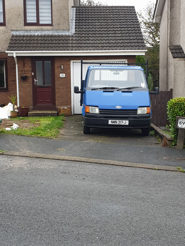 Street-level photograph showing a blue van parked on a paved driveway in front of a semi-detached house with a garage.