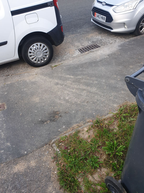 A close-up photograph of a paved driveway surface showing a white van and silver car, a drain grate, and a grass verge.