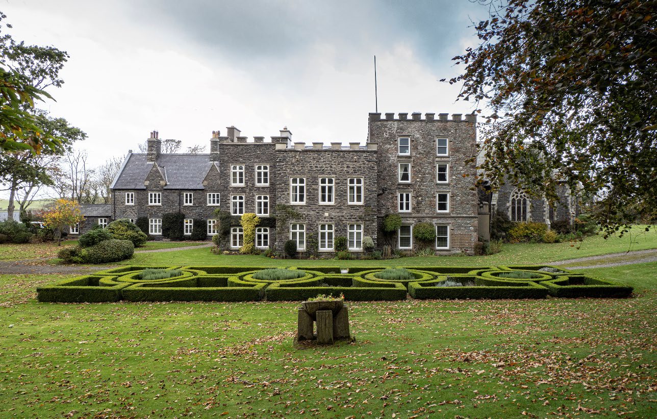 A wide exterior photograph of a large, historic grey stone manor house featuring crenellated rooflines and formal box hedge gardens in the foreground.