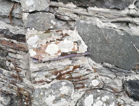 Close-up photograph of weathered layered stone masonry with lichen growth and a distinct repair patch.