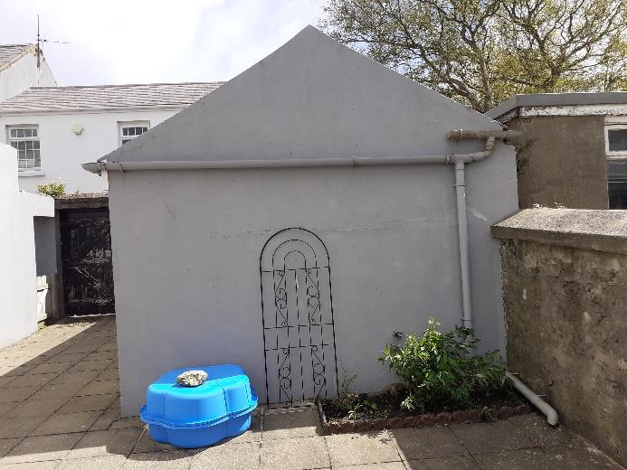 A photograph showing the front exterior of a small, grey single-story outbuilding or garage with a gable roof and a decorative metal screen. A blue plastic tub sits on the paved area in front of the structure.