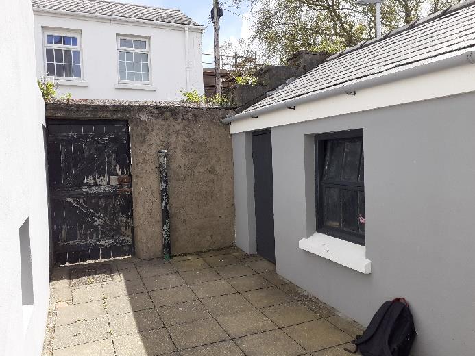 A photograph showing a paved courtyard area between a white residential building and a grey single-story outbuilding with a weathered gate nearby.
