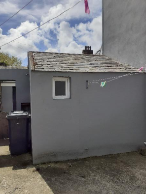A photograph showing a small, single-story grey building with a slate roof and a small window. A clothesline is strung across the front, and a wheelie bin is visible to the left.