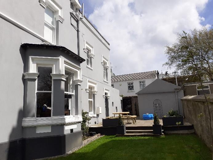A photograph showing the rear exterior of a white terraced building with a bay window, featuring a paved patio area, planters, and a small outbuilding in the background.