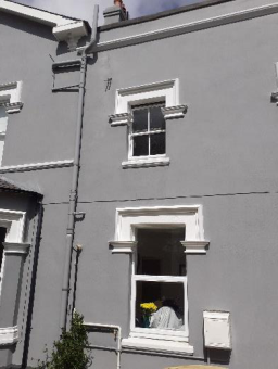 A close-up photograph of the grey exterior facade of a terraced building, showing white window frames, a drainpipe, and a small plant at the base.