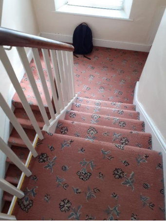 A photograph showing an interior staircase with pink floral carpet, white balusters, and a wooden handrail leading to a window.