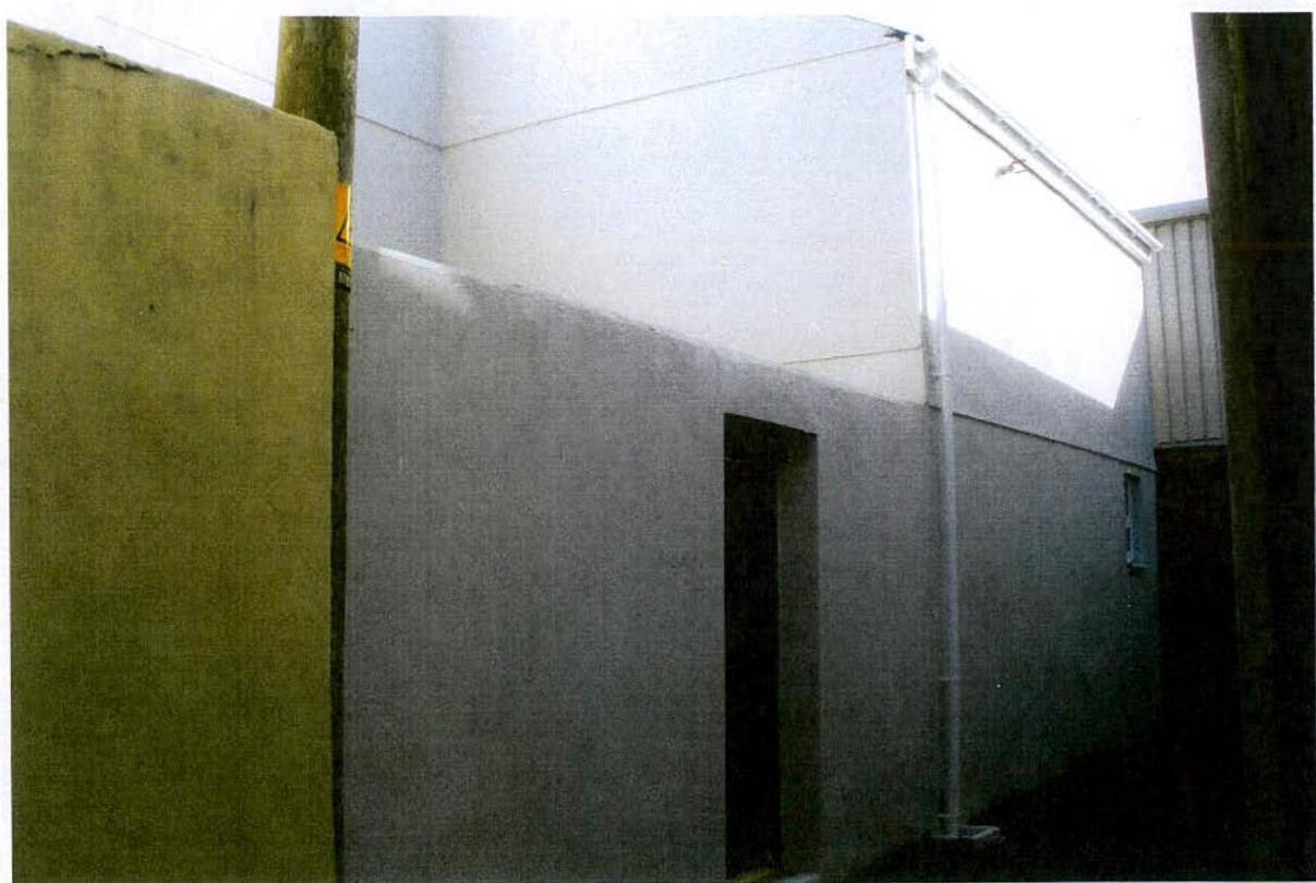 A grainy photograph showing the grey rendered exterior wall of a building with a dark doorway and a white downpipe running vertically.