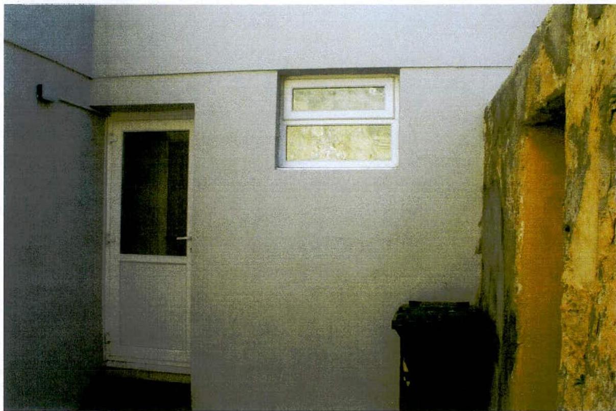 A photograph showing the exterior side elevation of a building featuring a white door and window next to a rough stone wall.