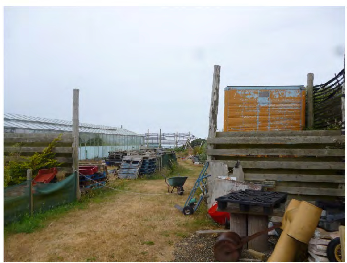 A photograph of a rural agricultural site featuring a long greenhouse structure on the left and a wooden fence with a rusty container on the right, surrounded by scattered farm equipment and pallets.