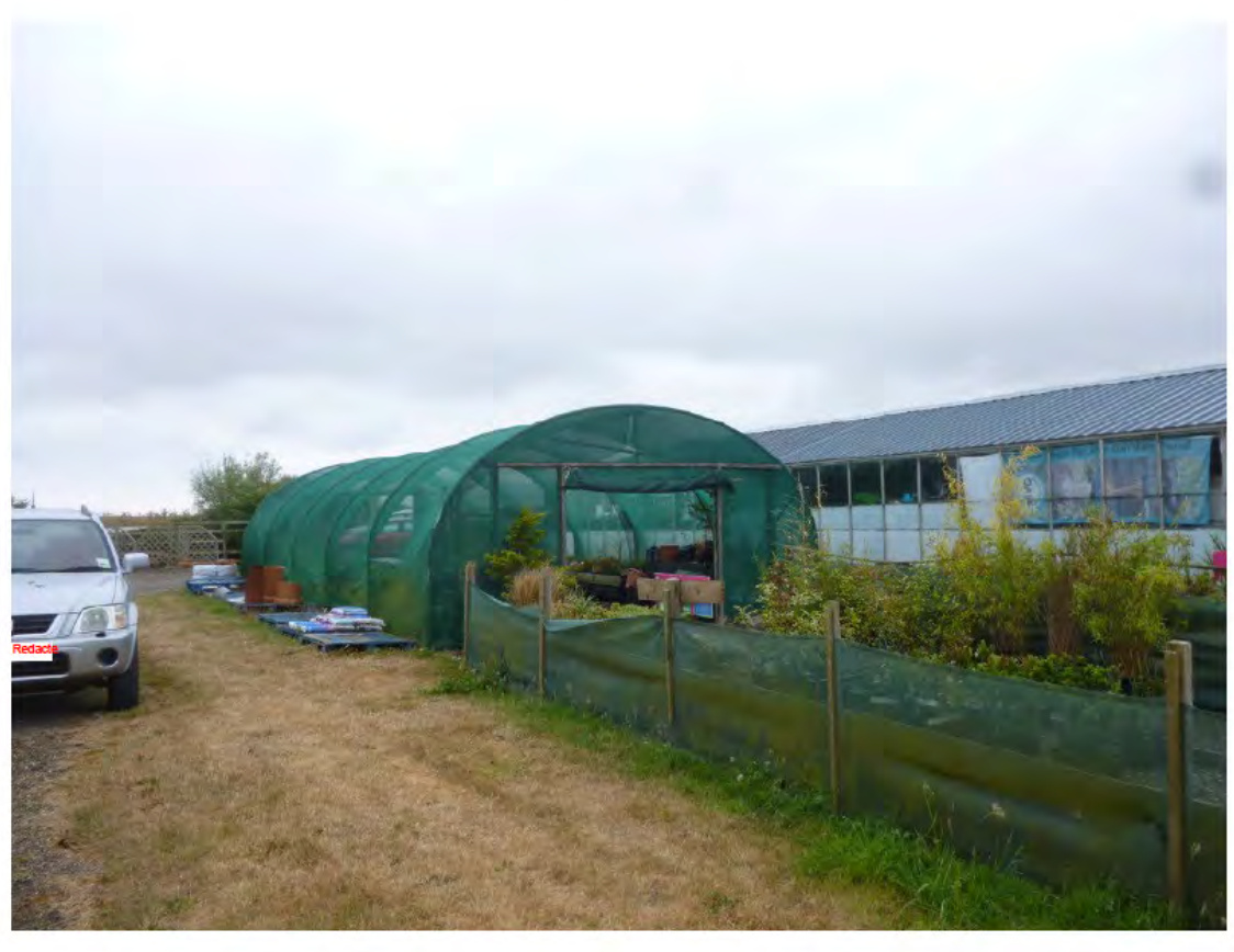 A photograph showing a rural site featuring a large green polytunnel and an adjacent long greenhouse structure with a metal roof.