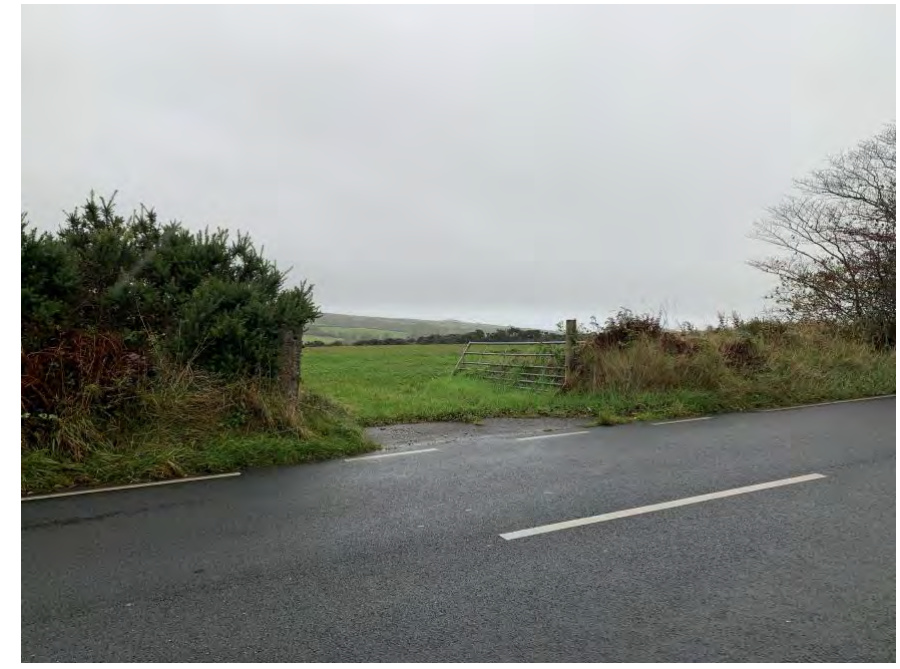 A photograph showing a rural road with a farm gate and track leading into a green field, likely the existing access point mentioned in the application.