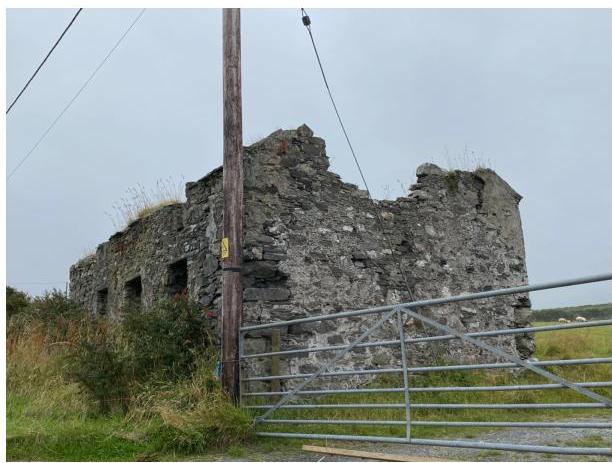 A photograph showing the dilapidated stone ruins of a traditional tholtan cottage with crumbling walls and no roof, viewed from behind a metal gate.