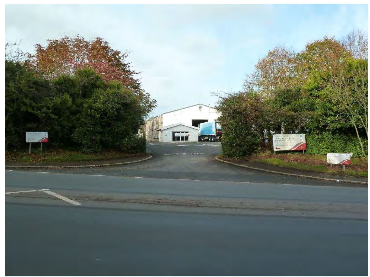 A street-level photograph showing the entrance to an industrial site with a white warehouse building and a truck, flanked by trees and entrance signs.