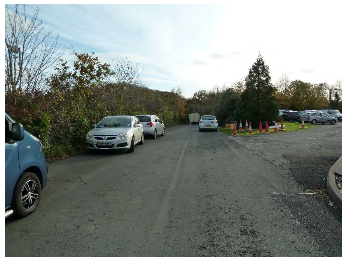 A photograph showing a paved access road and parking area with several cars parked on either side and orange traffic cones.