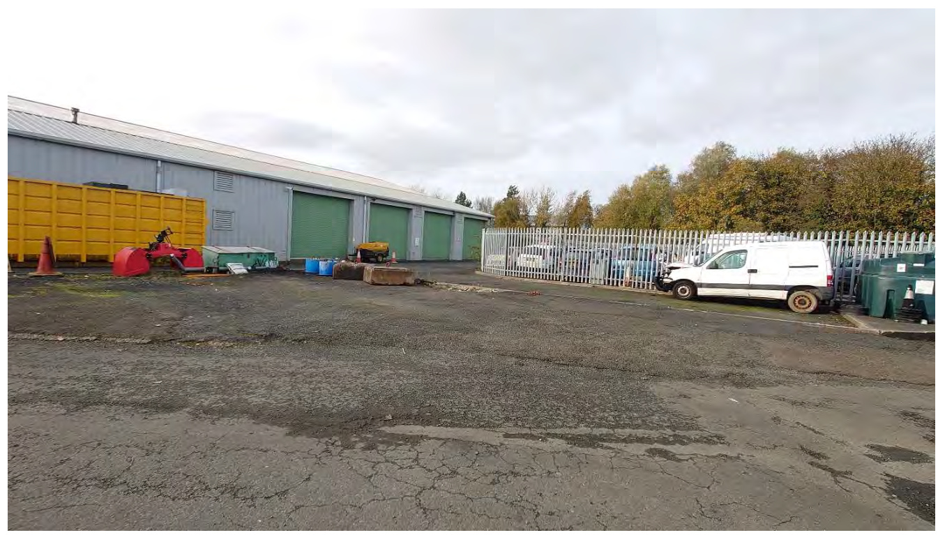 A photograph showing the exterior of an existing single-story industrial building with green roller shutter doors and a paved yard area.