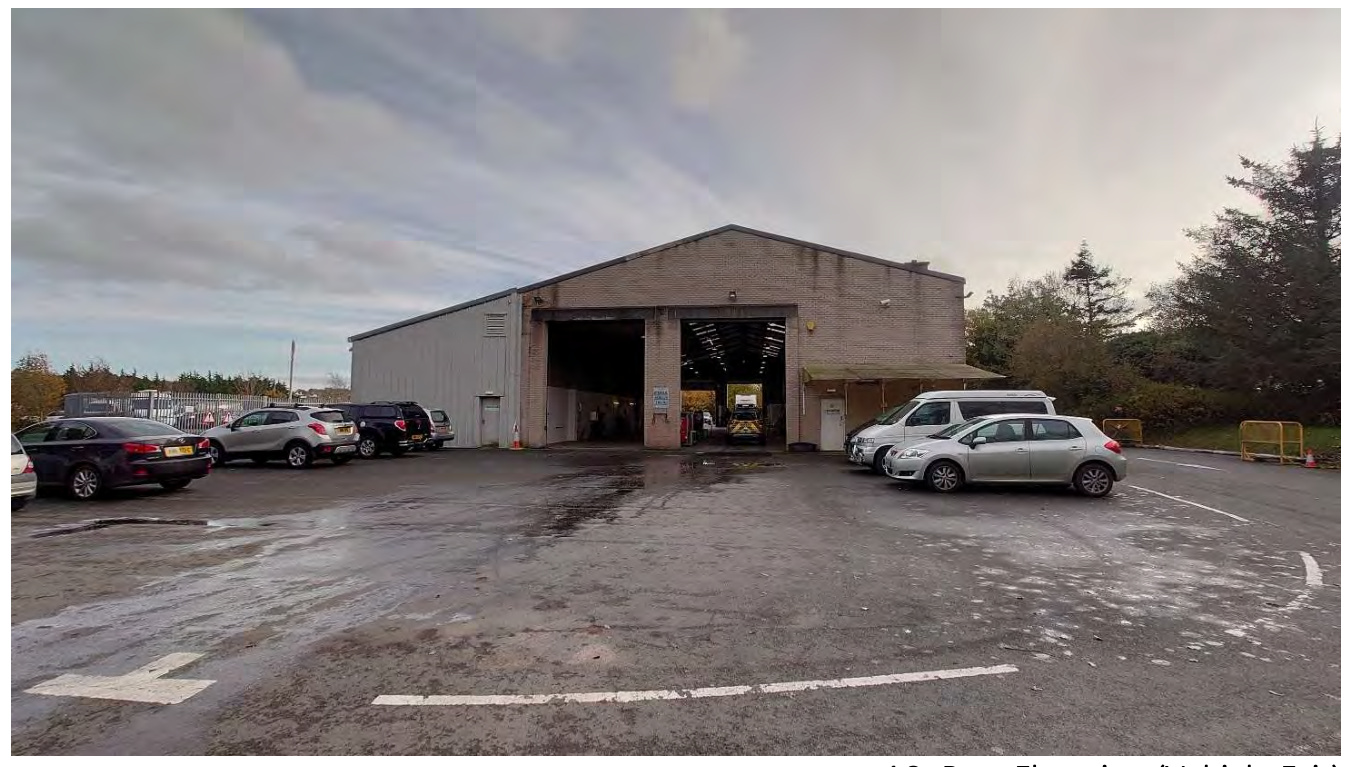 A photograph showing an existing single-story industrial building, identified as a vehicle test centre, with a paved parking area in the foreground containing several parked cars.