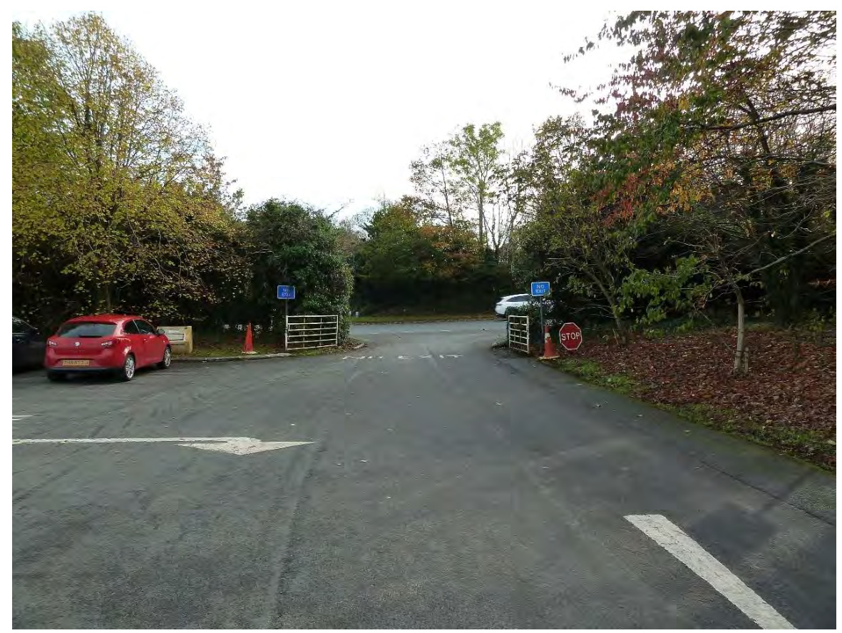 A photograph showing a paved entrance area with parked cars, trees, and traffic signage including a stop sign.