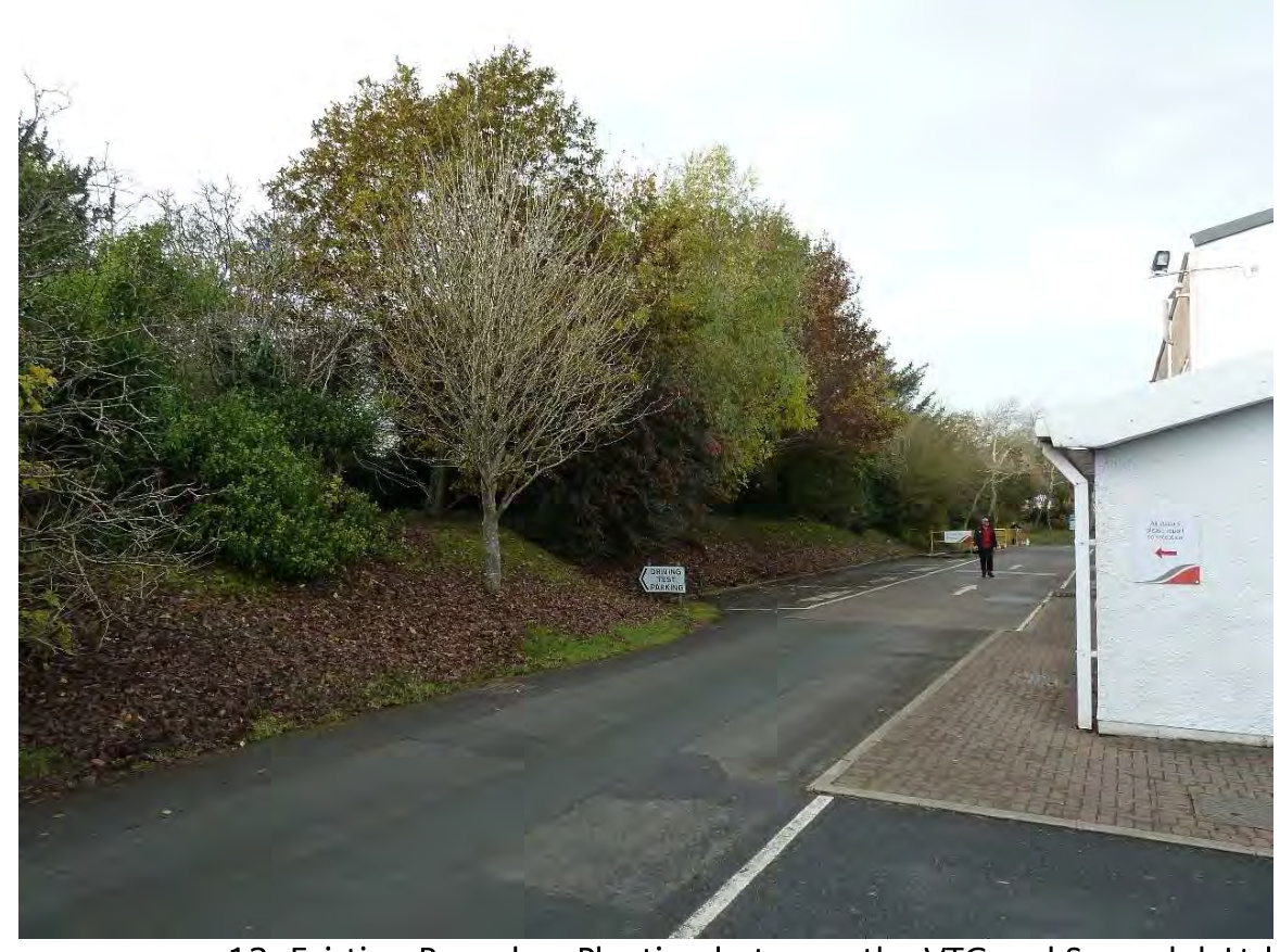 A street-level photograph showing a paved access road leading past a grassy bank with trees and a white building on the right, with a sign reading 'DRIVING TEST PARKED'.