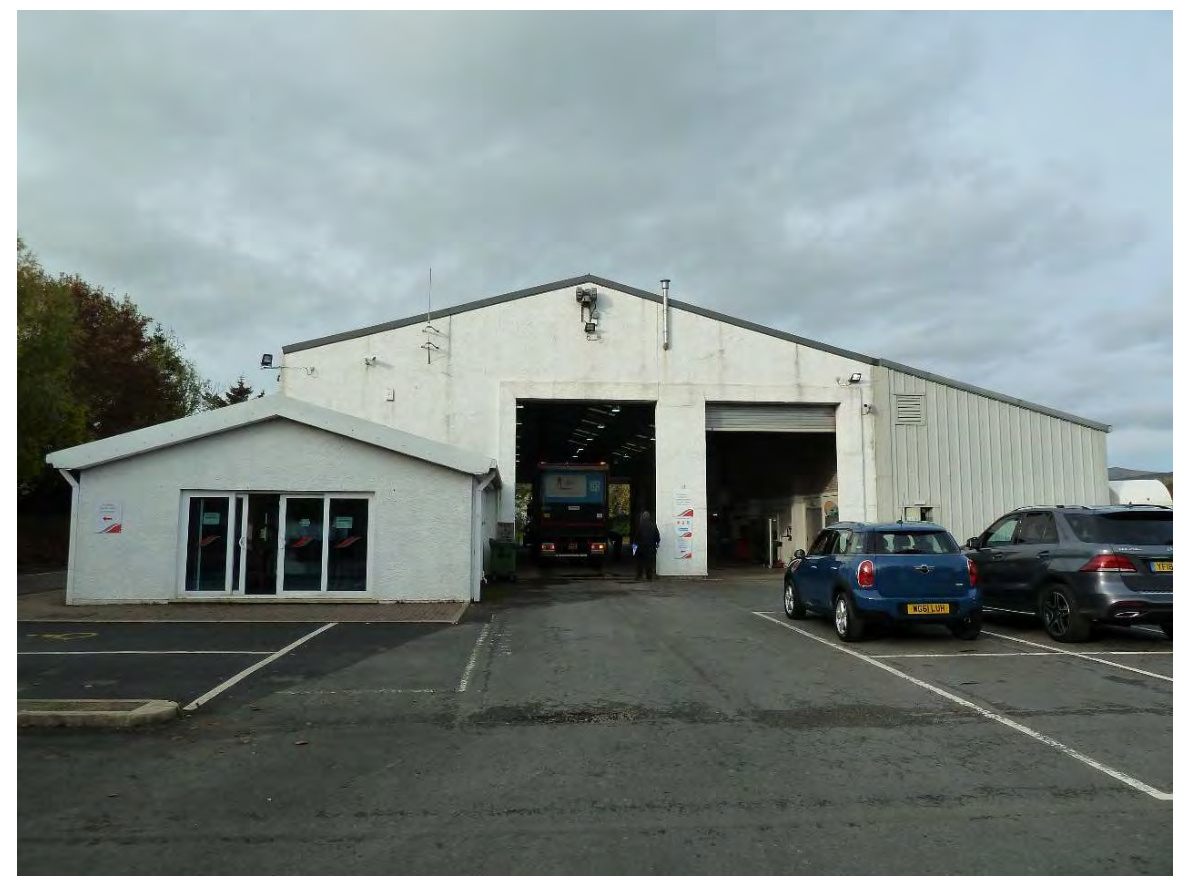 A photograph of the existing vehicle test centre, featuring a large white industrial building with open bays and an attached office section, with cars parked in the foreground.