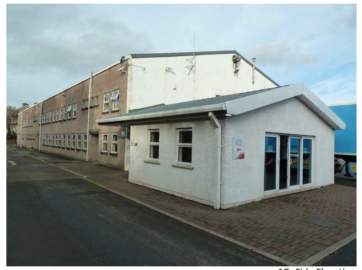 A photograph of the existing vehicle test centre, showing a long two-story brick building and an attached single-story white extension with a paved forecourt.