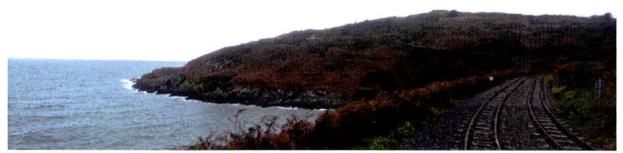 A panoramic photograph showing a coastal landscape with a railway track running along the base of a grassy hill next to the sea.