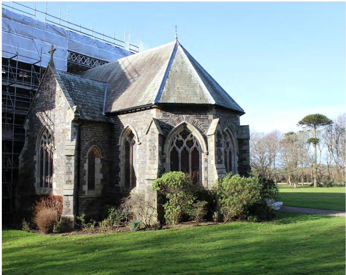 A photograph showing the exterior of a stone chapel with Gothic-style windows and scaffolding on the left side, set against a grassy lawn.