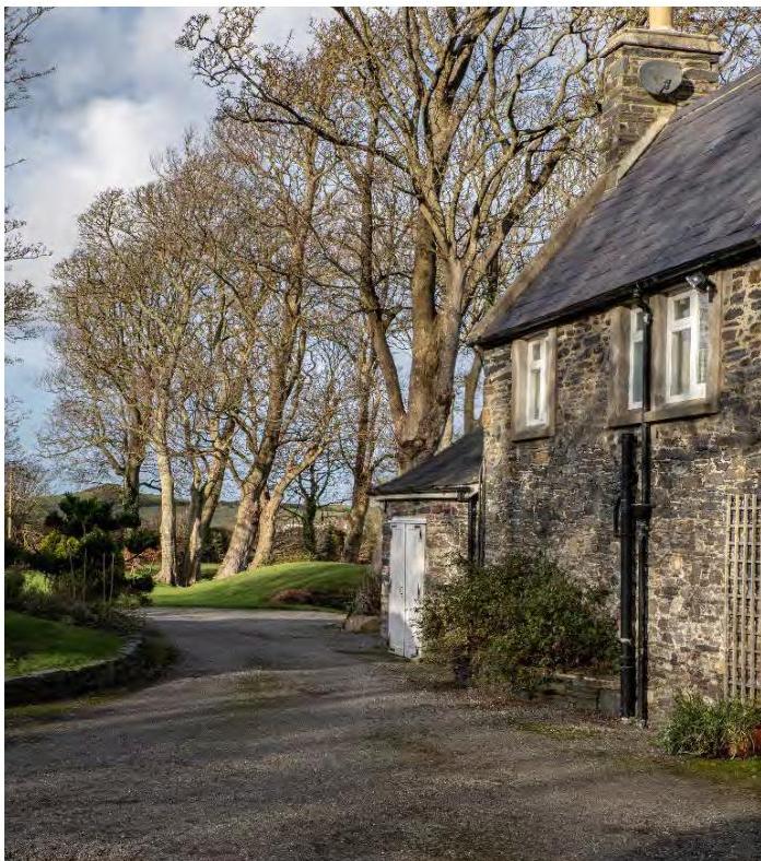 A photograph showing the exterior of a stone building with a slate roof, situated next to a gravel driveway and large trees.
