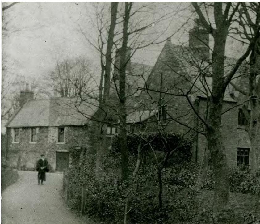 A black and white photograph showing a historic stone building complex with a person walking on a path in the foreground.