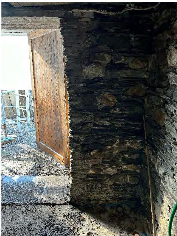 A close-up photograph showing a rough stone wall and a wooden door, likely part of the historic Chapel of St. Nicholas mentioned in the application.