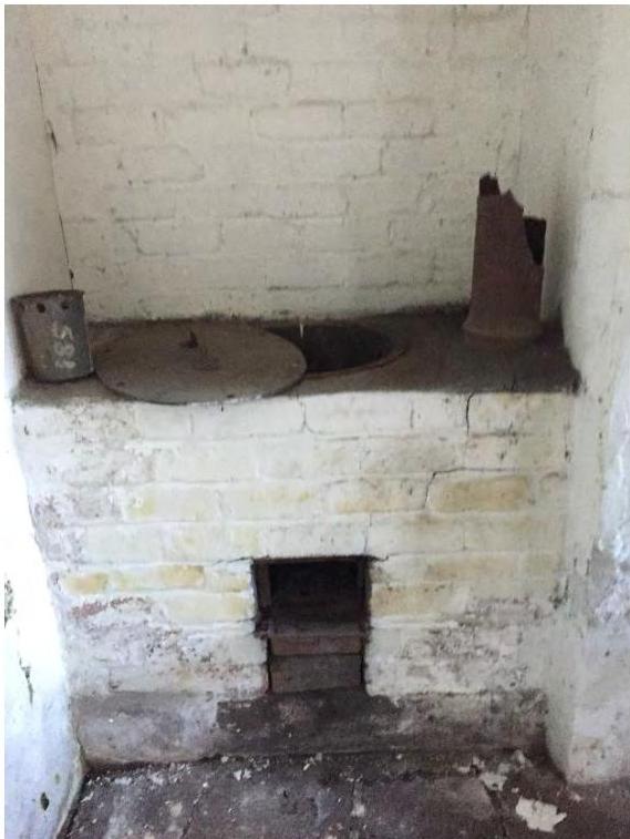 Interior photograph of an old white-washed brick range cooker or fireplace featuring a metal lid and rusty flue pipe.