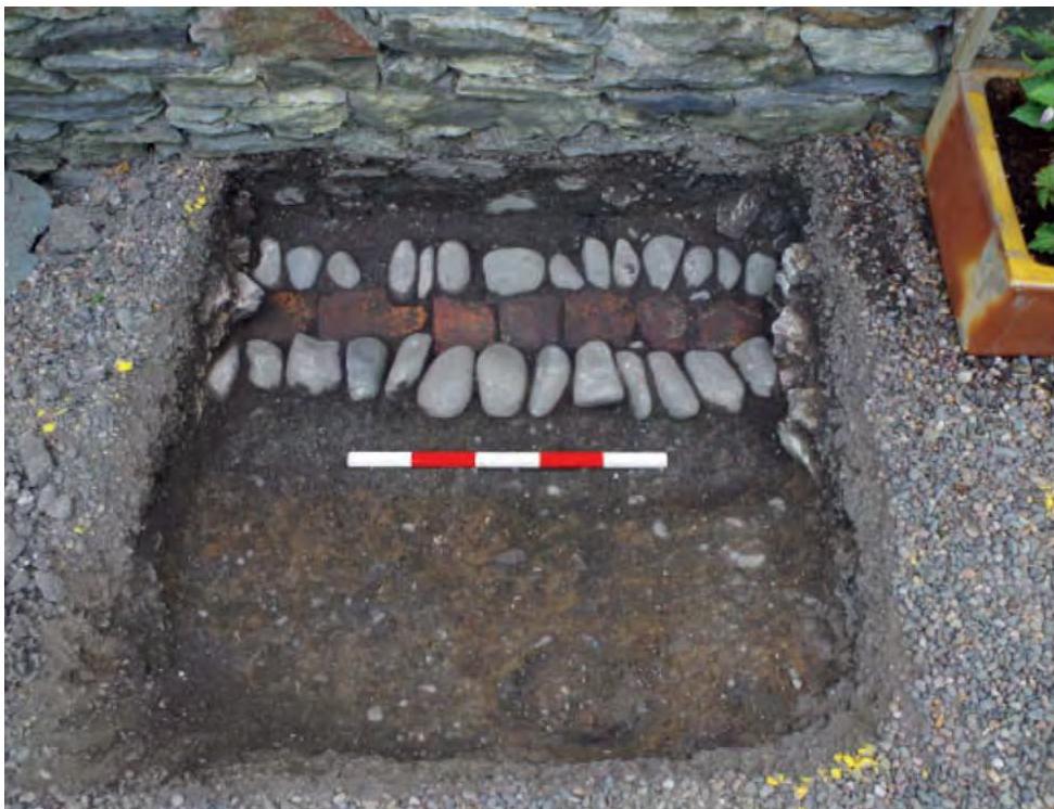 A photograph showing a square excavation pit dug next to a stone wall, revealing layers of stones and bricks with a red and white scale bar for reference.