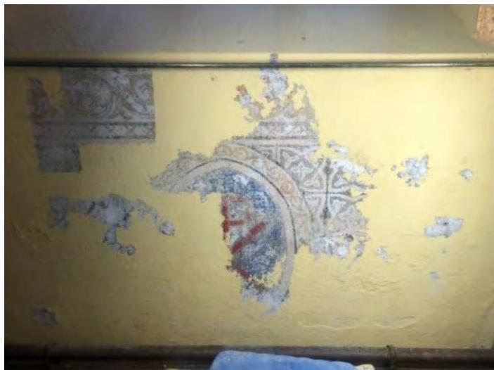 A close-up photograph of a yellow interior wall with peeling paint and exposed underlying decorative patterns, likely documenting the condition of a historic building.