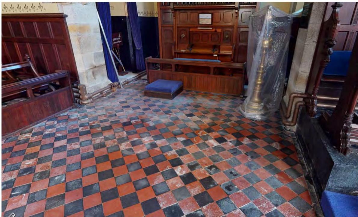Interior photograph of a historic chapel showing a red and black checkered tile floor, wooden pews, and stone walls with visible piping near the base.