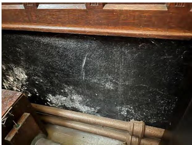 A close-up photograph of a dark, textured stone wall showing signs of damp or soot, with wooden pipes and a wooden structure in the foreground.