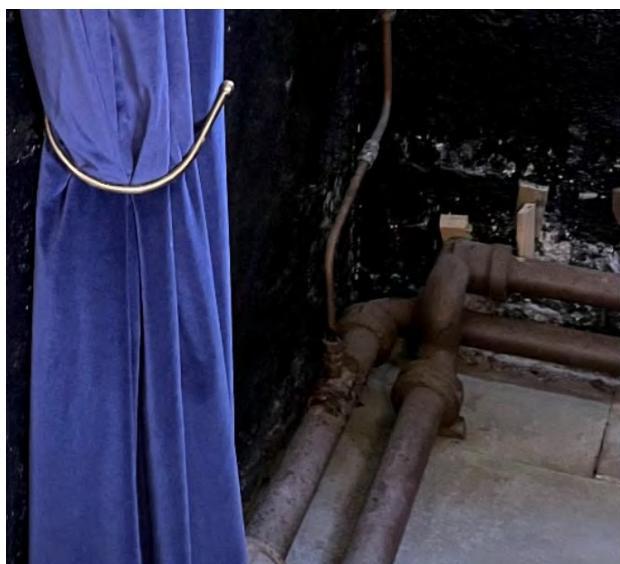 A close-up photograph showing rusty metal pipes running along a dark wall next to a blue curtain with a metal tie-back.