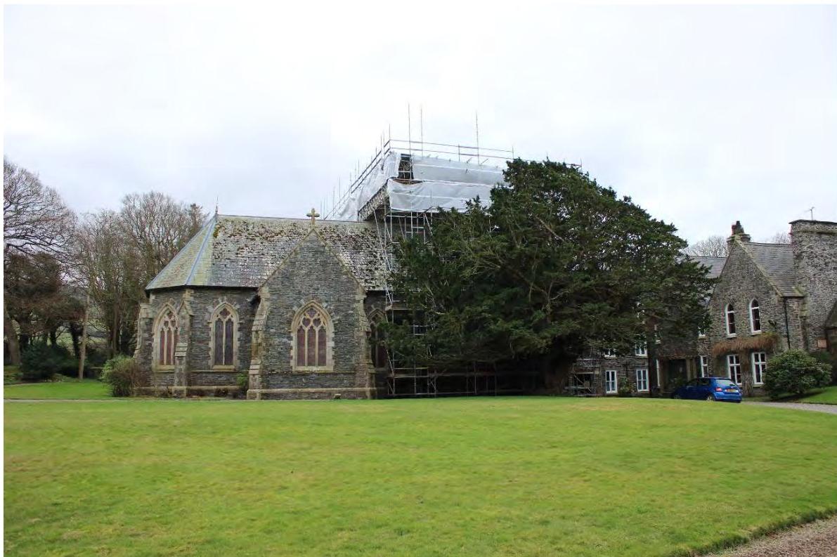 A photograph showing a stone chapel with Gothic windows adjacent to a larger stone building covered in scaffolding, set on a grassy lawn.