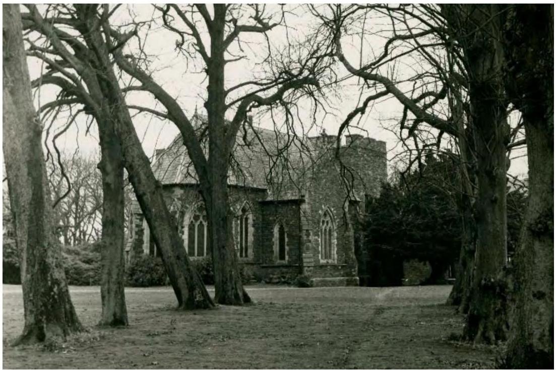 A black and white photograph showing a stone chapel or church building with Gothic-style windows viewed through large bare trees.