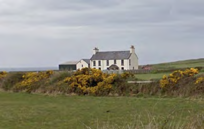 A photograph showing a white detached house with a grey roof situated in a rural landscape with yellow gorse bushes in the foreground.