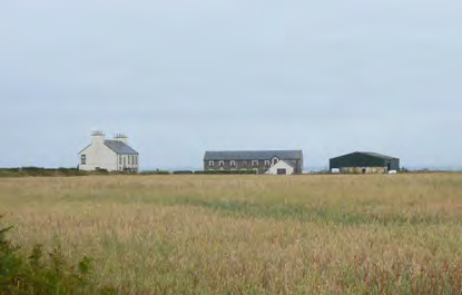 A photograph showing a rural landscape with a field in the foreground and three buildings in the distance, including a white house and agricultural structures.