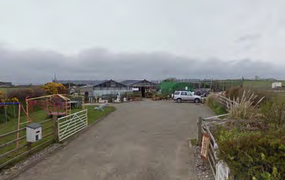 A photograph showing the existing site conditions of a rural property with a driveway leading to outbuildings and a garden area on the left.