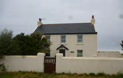 A photograph of a white, two-story detached house with a slate roof and chimneys. The property is situated behind a white boundary wall with a wooden gate.