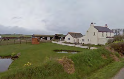 A photograph of a rural property featuring a large white detached house, smaller outbuildings, and a long barn-like structure in the background with a grassy foreground.