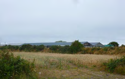 A photograph showing a grassy field in the foreground with existing buildings, including a large agricultural shed and a house, visible in the background under an overcast sky.