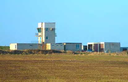 A photograph showing a grassy field in the foreground with a large, multi-story concrete building complex in the background under a clear blue sky.