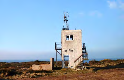 A photograph showing a small concrete tower structure with an external metal staircase situated on a grassy coastal landscape with the sea in the background.