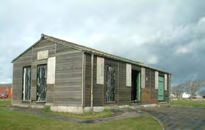 A photograph of an existing long, single-story wooden outbuilding or barn with boarded windows and green doors situated on a rural plot.