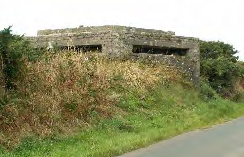 A photograph showing a concrete military pillbox or bunker situated on a grassy embankment next to a road.