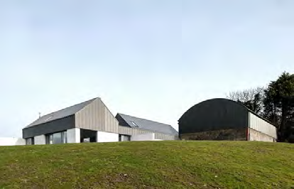 A photograph showing a grassy hillside with a modern pitched-roof building and a large curved agricultural shed in the background.