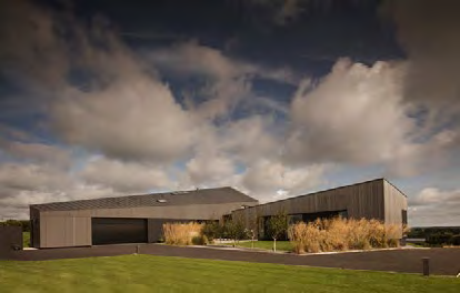 A photorealistic rendering of a modern, single-story detached house with an integral garage, featuring dark cladding and landscaped grounds.