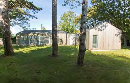 A photograph showing a grassy plot with mature trees in the foreground and existing outbuildings, including a wooden shed and greenhouse, in the background.